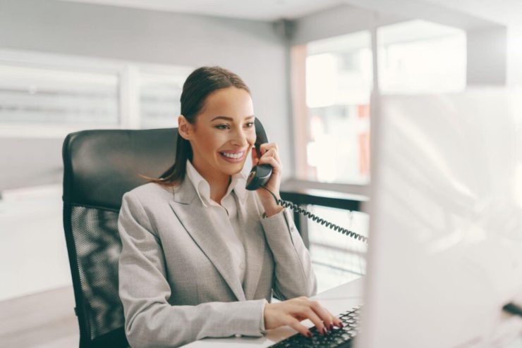 Smiling executive assistant in a gray blazer on a desk phone while typing at a computer in a bright office