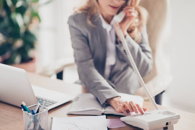 Executive assistant in a gray blazer dialing a desk phone while holding a planner open at an office desk