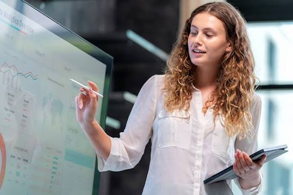 Project coordination assistant presenting a project analytics dashboard on a screen while holding a tablet