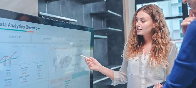 Project coordination assistant pointing to a data analytics dashboard on a large screen during a team meeting