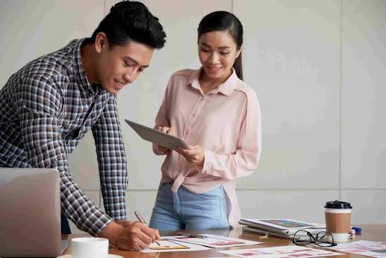Project coordinator writing up a project plan while a colleague checks the details on a tablet