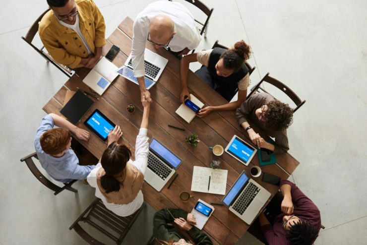 Overhead view of a project team around a wooden table with laptops and tablets as two colleagues shake hands