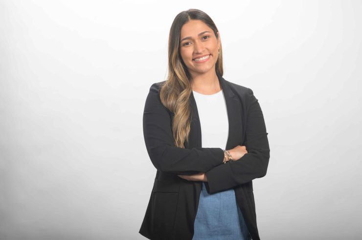 Smiling executive assistant with long hair in a black blazer and white top, arms crossed, against a light gray studio background