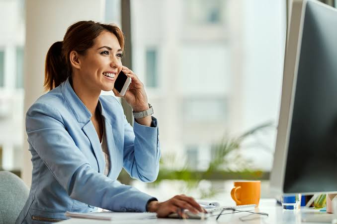 Smiling professional in a light blue blazer on a phone call while working at a computer in a bright office
