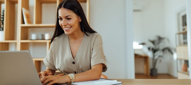 Smiling executive research assistant typing on a laptop at a wooden desk in a bright home office