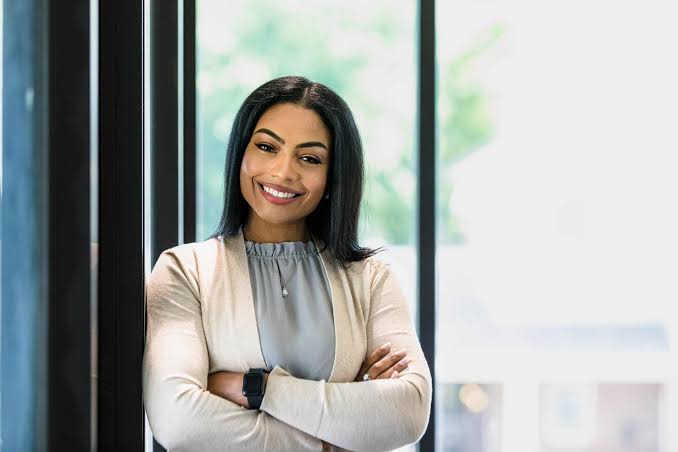 Smiling executive assistant in a cream cardigan and gray blouse, arms crossed, standing beside an office window